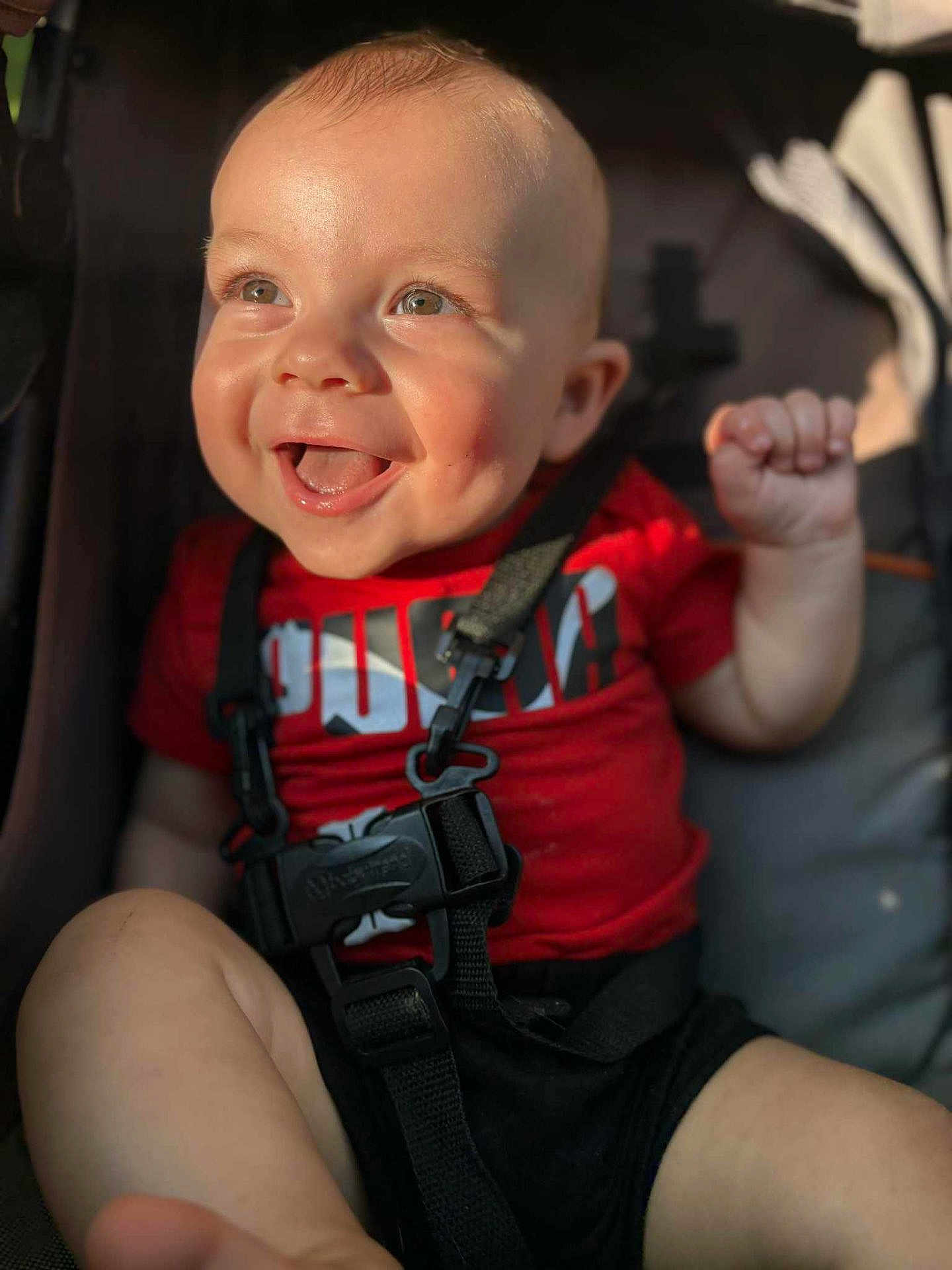 Cooper is registered to the contest to win money with this photo: baby, child, smiling, happy, face, red_shirt, stroller, safety_strap, sunlight, dimples, skin, hand, foot, infant, cute, portrait, closeup, daylight, person, outdoor