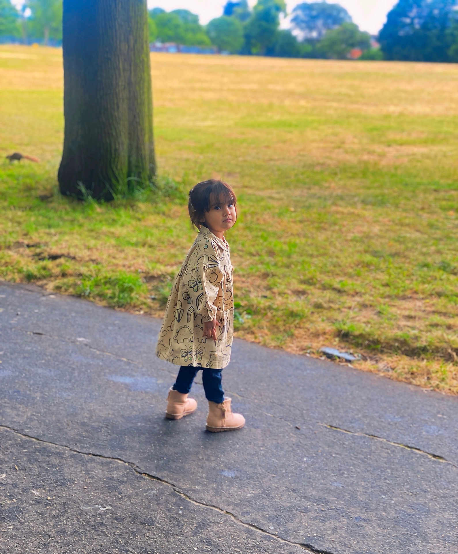 Airah is registered to the contest to win money with this photo: child, park, coat, dinosaur_pattern, butterfly_pattern, boots, paved_path, grass, tree, outdoor, nature, curious, young_child, casual_clothing, walking, daylight, fall, scenery, person, portrait