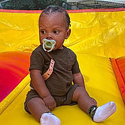 Kayden joined the competition — help win amazing prizes! child, toddler, pacifier, braided_hair, sitting, yellow_inflatable, outdoor, play_area, socks, shorts, tshirt, curious, baby, summer, fun, bright_colors, daytime, trees, tent, playground