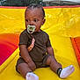 child, toddler, pacifier, braided_hair, sitting, yellow_inflatable, outdoor, play_area, socks, shorts, tshirt, curious, baby, summer, fun, bright_colors, daytime, trees, tent, playground