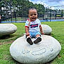 Kayden is registered to the contest to win money with this photo: child, clouds, daytime, fence, grass, happy, nature, outdoor, park, playful, rock, shoes, shorts, sitting, sky, smiling, tennis_court, toddler, trees, tshirt