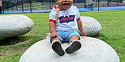 Kayden joined the competition — help win amazing prizes! child, clouds, daytime, fence, grass, happy, jeans, outdoor, park, playful, shoes, sitting, sky, smiling, stone, tennis_court, toddler, trees, tshirt, young_child