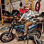 Aiden is registered to the contest to win money with this photo: child, boy, dirt_bike, motorcycle, barefoot, smiling, curly_hair, indoor, garage, workshop, tools, scaffolding, hose, muddy, clothing, pants, shirt, footwear, person, vehicle
