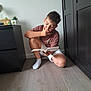 child, boy, reading, book, indoor, floor, crouching, thinking, casual_clothing, shorts, socks, furniture, cabinet, drawer, plant, home, concentration, light, wooden_floor, young