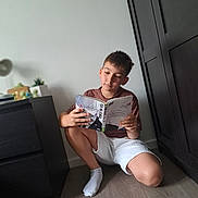 Eladio participe au concours pour gagner de l'argent avec cette photo : boy, child, reading, book, indoor, floor, furniture, cabinet, sock, shorts, tshirt, concentration, portrait, home, casual, young, brown_hair, light, shadow, relaxation