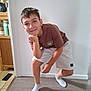 boy, child, indoor, pose, smile, shorts, tshirt, socks, floor, wall, furniture, shelf, cabinet, light_switch, wood_floor, casual_clothing, happy, person, home, portrait