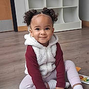 Harlow joined the competition — help win amazing prizes! child, toddler, portrait, smiling, sitting, indoors, floor, wood_floor, shelving, toy, clothing, fleece_vest, hood, hair_buns, curly_hair, pink_pants, socks, happy, camera_facing, home