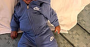 Kiari is registered to the contest to win money with this photo: baby, child, infant, nike, tracksuit, blue_clothing, socks, couch, pillow, indoor, person, face, smile, relaxing, young_child, curious, seated, furniture, home, casual