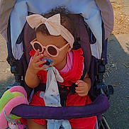 Orabella a rejoint le concours — aidez-le/la à gagner de superbes lots ! toddler, stroller, sunglasses, headband, pacifier, toy, outdoor, child, footwear, red_dress, baby, sunlight, seat, cute, accessory, summer, person, walking, shade, sidewalk