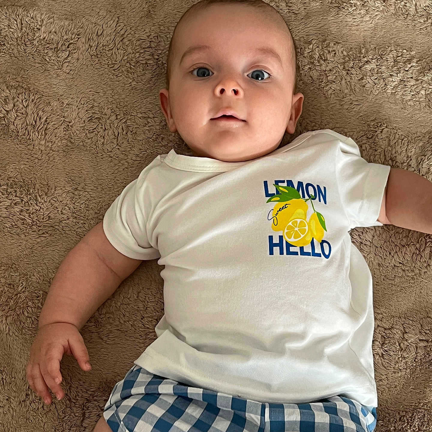 élio a rejoint le concours — aidez-le/la à gagner de superbes lots ! baby, blue_shorts, carpet, casual_clothing, checkered, child, cute, expression, eyes_open, floor, hand, indoor, infant, lemon, lying_down, person, portrait, skin, tshirt, young
