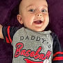 élio a rejoint le concours — aidez-le/la à gagner de superbes lots ! baby, baseball, casual, child, closeup, clothing, cute, face, happy, indoor, infant, lying_down, person, portrait, purple_blanket, shirt, smiling, sports, text, young_child