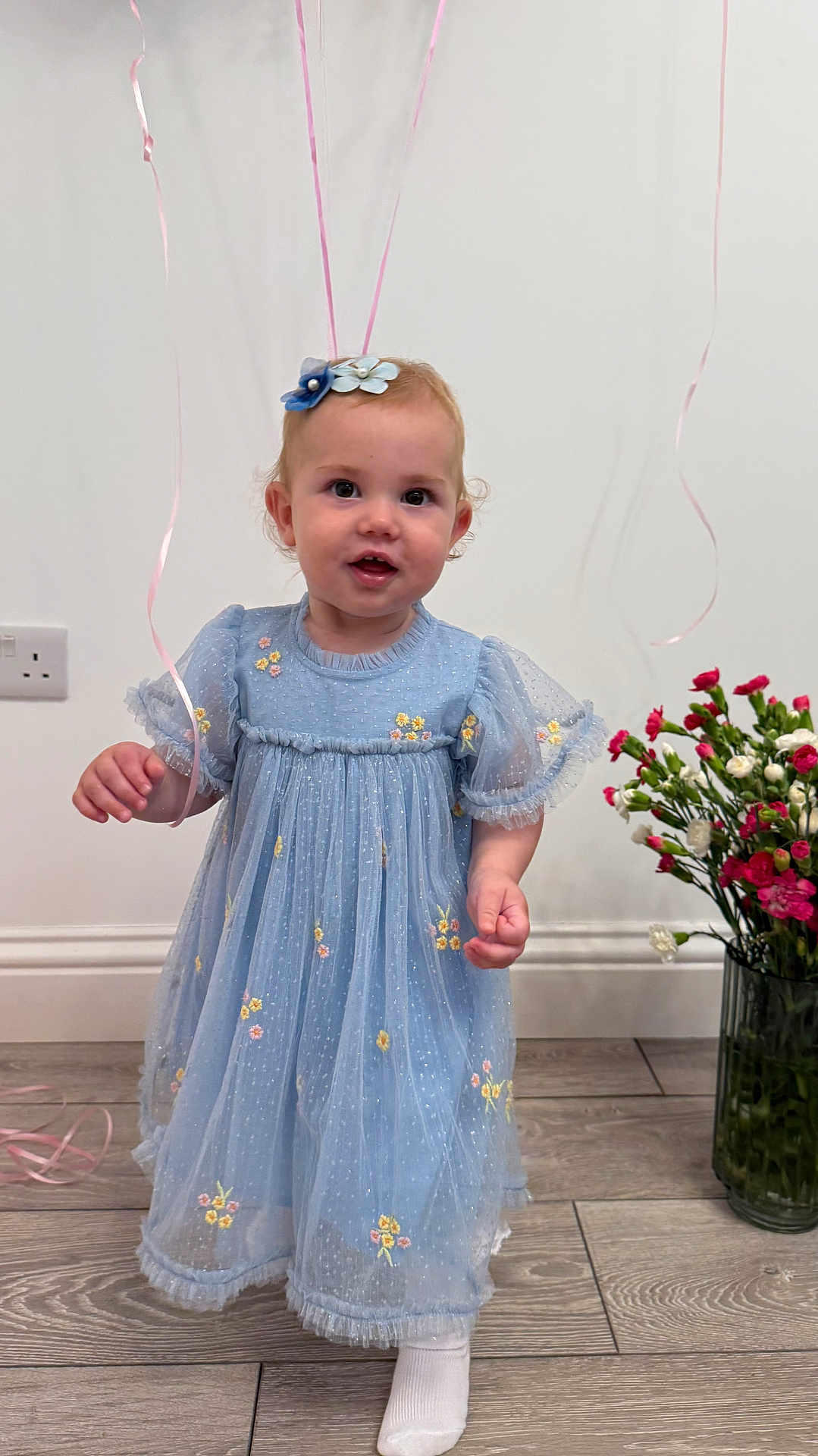 Agnes is registered to the contest to win money with this photo: toddler, child, blue_dress, balloons, flowers, vase, wooden_floor, indoor, smiling, cute, portrait, person, celebration, decor, happy, flower_bow, socks, curly_hair, standing, young_child