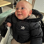 Maylon participe au concours pour gagner de l'argent avec cette photo : baby, child, stroller, puffer_jacket, black_jacket, adidas_logo, rosy_cheeks, blue_eyes, indoor, radiator, shoe_shelf, shoes, blanket, tile_floor, table, hood, hand, pram, portrait, cozy