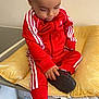 toddler, child, red_tracksuit, sneakers, yellow_cushion, indoor, drawer, household_items, black_shoes, sitting, curious, baby, clothing, footwear, person, casual, young_child, wall, table, home