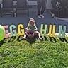child, toddler, grass, basket, eggs, egg_balloon, letters, egg_hunt, outdoor, sunlight, shadow, greenery, sidewalk, patio_chairs, person, playful, fun, spring, holiday, celebration