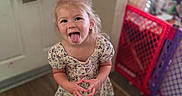 Astrid joined the competition — help win amazing prizes! child, girl, tongue_out, pink_bow, floral_dress, brown_boots, wooden_floor, baby_gate, indoor, door, towel, playful, smiling, person, home, cute, blonde_hair, standing, expression, fun