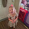 child, girl, tongue_out, pink_bow, floral_dress, brown_boots, wooden_floor, baby_gate, indoor, door, towel, playful, smiling, person, home, cute, blonde_hair, standing, expression, fun