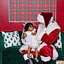 child, toddler, santa_claus, christmas, holiday, festive, dress, bow, curly_hair, smile, green_couch, pillows, plaid_background, red_wall, boots, indoor, celebration, happy, person, family