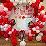 child, toddler, birthday, balloons, strawberry, banner, party_decor, balloon_arch, red_balloons, pink_balloons, white_balloons, number_one_balloon, wood_floor, sitting, dress, hair_bow, celebration, decorations, smile, infant