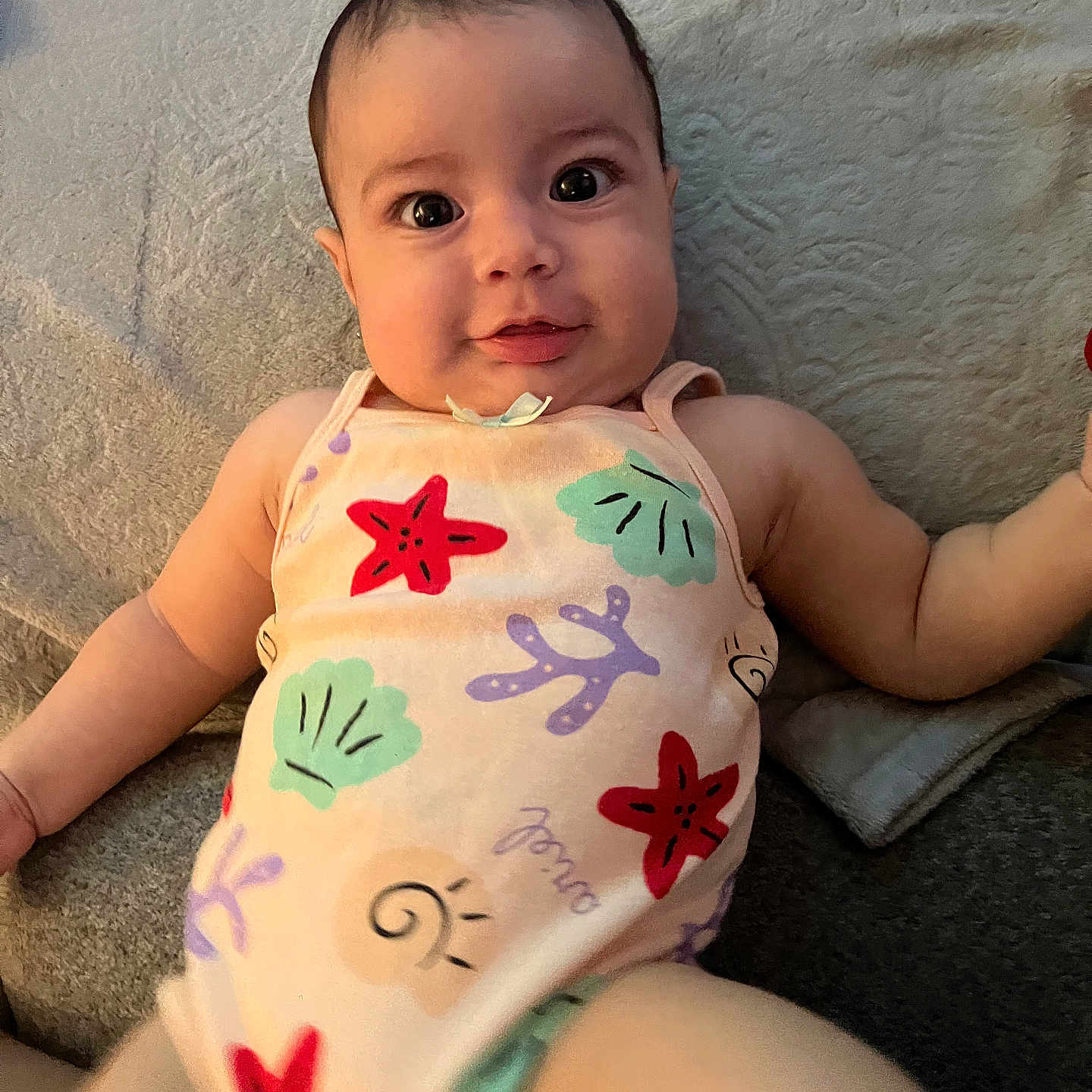 Aria joined the competition — help win amazing prizes! baby, child, infant, swimsuit, colorful_clothing, lying_down, blanket, face, eyes, smile, skin, cute, playful, indoors, person, young_child, expression, summer_wear, patterned_clothing, happy