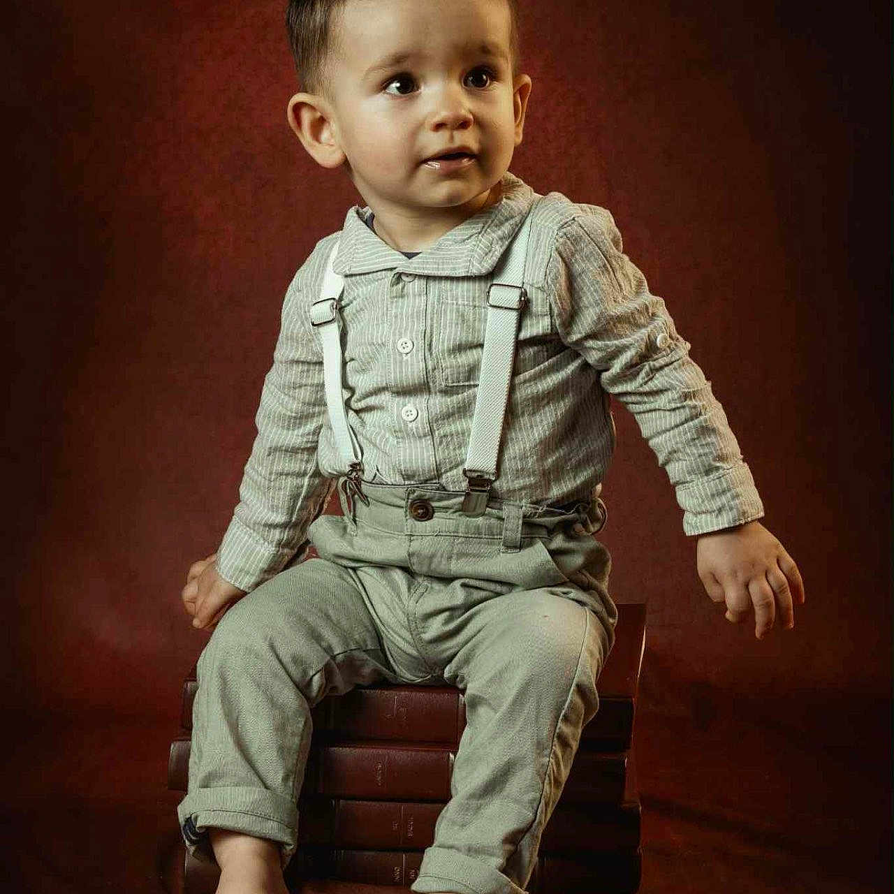 Mélyo a rejoint le concours — aidez-le/la à gagner de superbes lots ! barefoot, books, boy, brown_background, child, clothing, curious, cute, expression, fashion, indoors, person, photography, portrait, sitting, stack, studio, suspenders, toddler, young_child