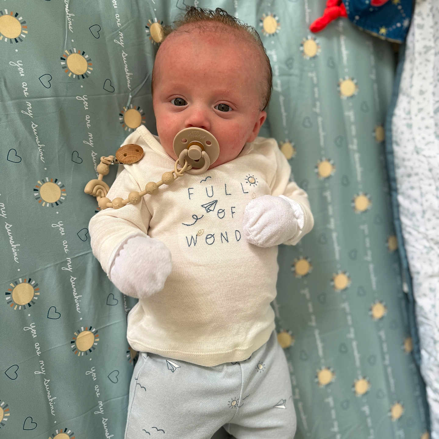 Nolan is registered to the contest to win money with this photo: baby, blanket, blue, clothing, comfort, cute, expression, face, heart, indoors, infant, lying_down, mittens, pacifier, pattern, small_hands, soft_toys, sun, white, wooden_clip