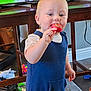 toddler, child, lollipop, blue_outfit, indoor, carpet, toy, television, table, wooden_furniture, curious, standing, home, play_area, cute, baby, face, hand, room, floor