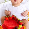 Jayden Berea is registered to the contest to win money with this photo: baby, child, infant, portrait, sitting, white_clothing, dark_hair, wide_eyes, serious_expression, fruit, watermelon, strawberries, grapes, oranges, fruit_cake, birthday, indoor, bed, close_up, food