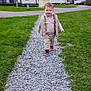 toddler, child, walking, gravel_path, grass, outdoor, shoes, sneakers, clothing, vest, houses, residential_area, daytime, sky, trees, smiling, person, casual, cute, young