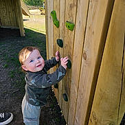 Romeo is registered to the contest to win money with this photo: toddler, child, playground, climbing_wall, wooden_structure, green_climbing_holds, outdoor, daylight, play, curious, blue_eyes, casual_clothing, jacket, pants, sock, shoe, grass, fence, smile, person