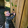 toddler, child, playground, climbing_wall, wooden_structure, green_climbing_holds, outdoor, daylight, play, curious, blue_eyes, casual_clothing, jacket, pants, sock, shoe, grass, fence, smile, person