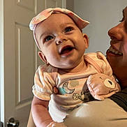 Octavia is registered to the contest to win money with this photo: baby, infant, smiling, headband, mittens, adult, person, indoor, door, clothing, holding, cute, happy, face, portrait, closeup, child, expression, parent, love