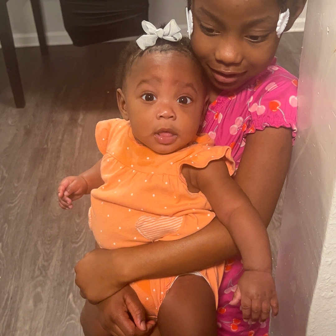 Skylnn is registered to the contest to win money with this photo: baby, child, curly_hair, family, floor, furniture, hair_bow, happy, holding, indoor, orange_clothing, person, pink_clothing, sitting, smile, snack_bag, table, toddler, wall, wood_floor