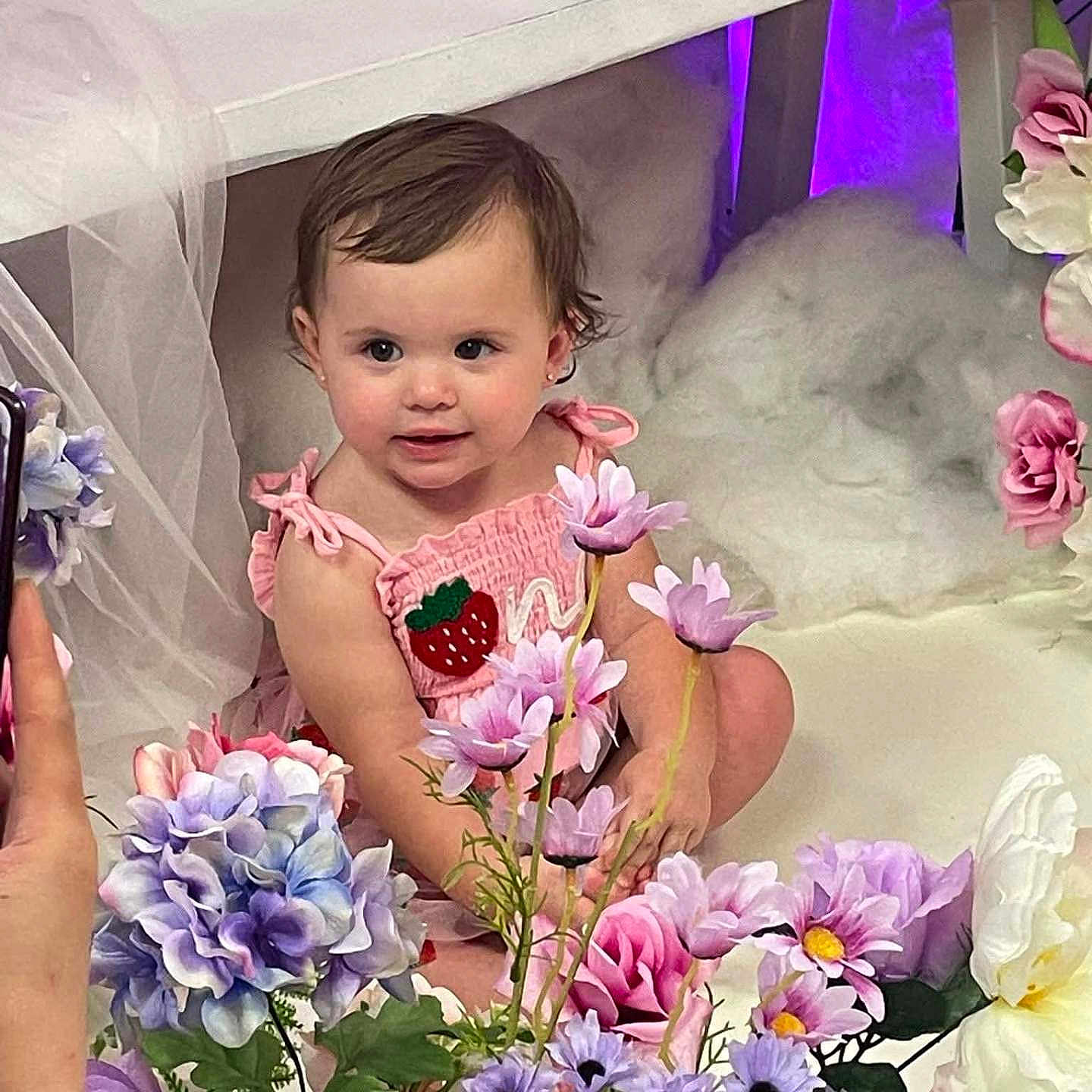 Vyolette is registered to the contest to win money with this photo: baby, child, cute, decor, flower_arrangement, hand, human, indoor, person, phone, photography, pink_dress, pink_flowers, portrait, purple_flowers, sitting, smiling, strawberry_patch, toddler, white_flowers