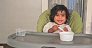 Kashif joined the competition — help win amazing prizes! toddler, high_chair, smiling, indoor, wooden_floor, wall_clock, wall_decal, spoon, bowl, child, happy, baby, feeding, green_cushion, plastic_container, home, doorway, curtains, flooring, person