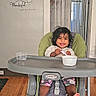 toddler, high_chair, smiling, indoor, wooden_floor, wall_clock, wall_decal, spoon, bowl, child, happy, baby, feeding, green_cushion, plastic_container, home, doorway, curtains, flooring, person