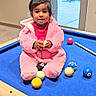 toddler, child, pink_outfit, pool_table, billiard_balls, indoor, glass_door, playing, happy, cute, cross_legged, carpeted_floor, wooden_frame, casual, young_child, smiling, curious, bright_lighting, modern_interior, fun