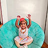 Kashif joined the competition — help win amazing prizes! toddler, child, girl, smiling, happy, sitting, chair, indoor, headband, flower, colorful_clothing, striped_leggings, shoes, wooden_floor, wall, necklace, cute, raising_hands, portrait, furniture