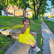 Kashif is registered to the contest to win money with this photo: child, toddler, yellow_dress, smiling, stone_wall, grass, tree, sidewalk, suburban, sunlight, outdoor, greenery, house, casual_clothing, sneakers, happy, person, daytime, nature, playful