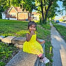 child, toddler, yellow_dress, smiling, stone_wall, grass, tree, sidewalk, suburban, sunlight, outdoor, greenery, house, casual_clothing, sneakers, happy, person, daytime, nature, playful