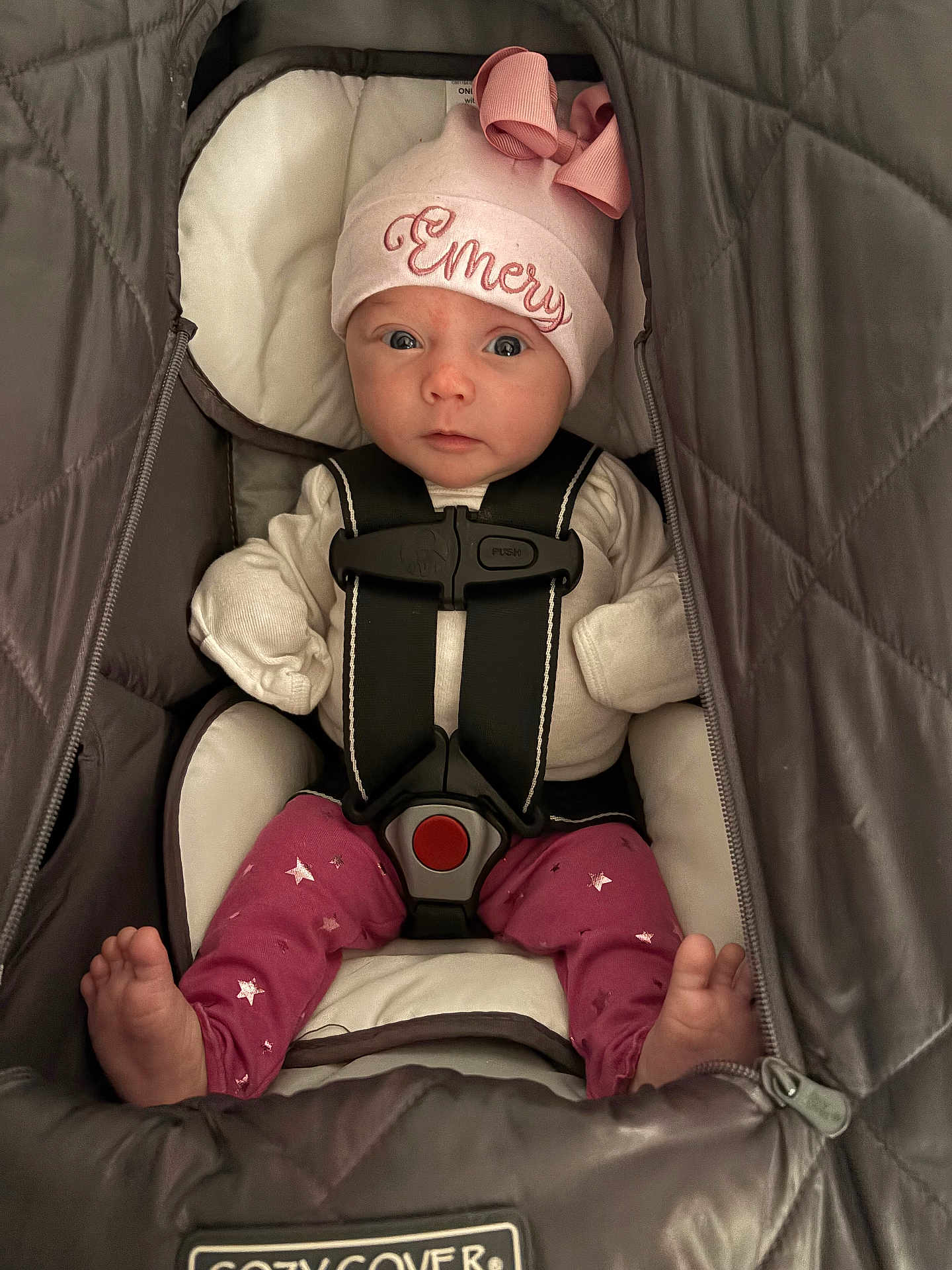 Emery is registered to the contest to win money with this photo: baby, infant, car_seat, pink_hat, bow, clothing, face, child, cute, looking_at_camera, safety_harness, foot, star_pattern, warm, indoor, person, adorable, newborn, seated, cozy