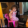 child, toddler, curly_hair, pink_chair, striped_dress, indoor, smiling, wooden_shelf, books, carpet, floor, room, furniture, person, playful, casual_clothing, sneakers, home, young_child, portrait
