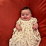 baby, infant, smile, tongue_out, dress, floral_pattern, cute, chubby_cheeks, sitting, blanket, cozy, child, portrait, happy, indoors, person, young_child, soft_texture, warm_colors, playful