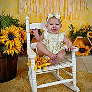 Mckinlyn is registered to the contest to win money with this photo: baby, child, smiling, rocking_chair, sunflower, headband, dress, flower, yellow, indoor, happy, cute, portrait, wooden_chair, floor_rug, decor, infant, seated, background, playful