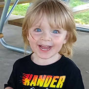 Xander is registered to the contest to win money with this photo: child, smiling, blue_eyes, blond_hair, toddler, black_shirt, text_on_shirt, outdoor, picnic_table, metal_legs, concrete_floor, green_background, happy, portrait, person, casual_clothing, young_child, daylight, excited, cute
