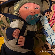 Kashton is registered to the contest to win money with this photo: baby, infant, pacifier, hoodie, hat, camo_pattern, blanket, leopard_print, yellow_quilt, clothing, cute, face, hand, person, indoors, lying_down, blue, green, newborn, child