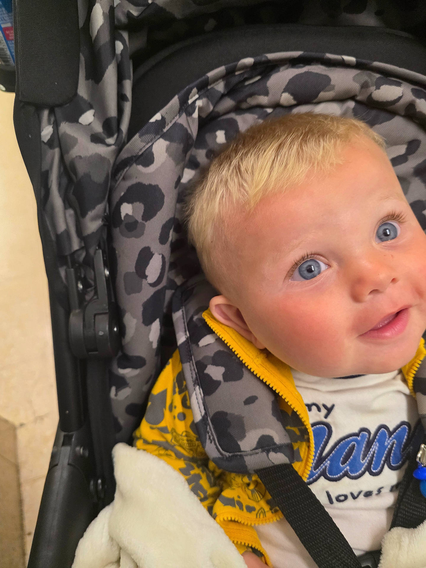 Parker is registered to the contest to win money with this photo: baby, blanket, blonde_hair, blue_eyes, child, clothing, curious, cute, face, happy, head, indoors, infant, leopard_print, patterned_fabric, person, seated, soft_texture, stroller, yellow_jacket