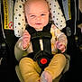 baby, car_seat, infant, winking, yellow_pants, patterned_shirt, socks, straps, safety_harness, cute, indoor, child, person, seat, head_support, playful, expression, smile, closeup, adorable