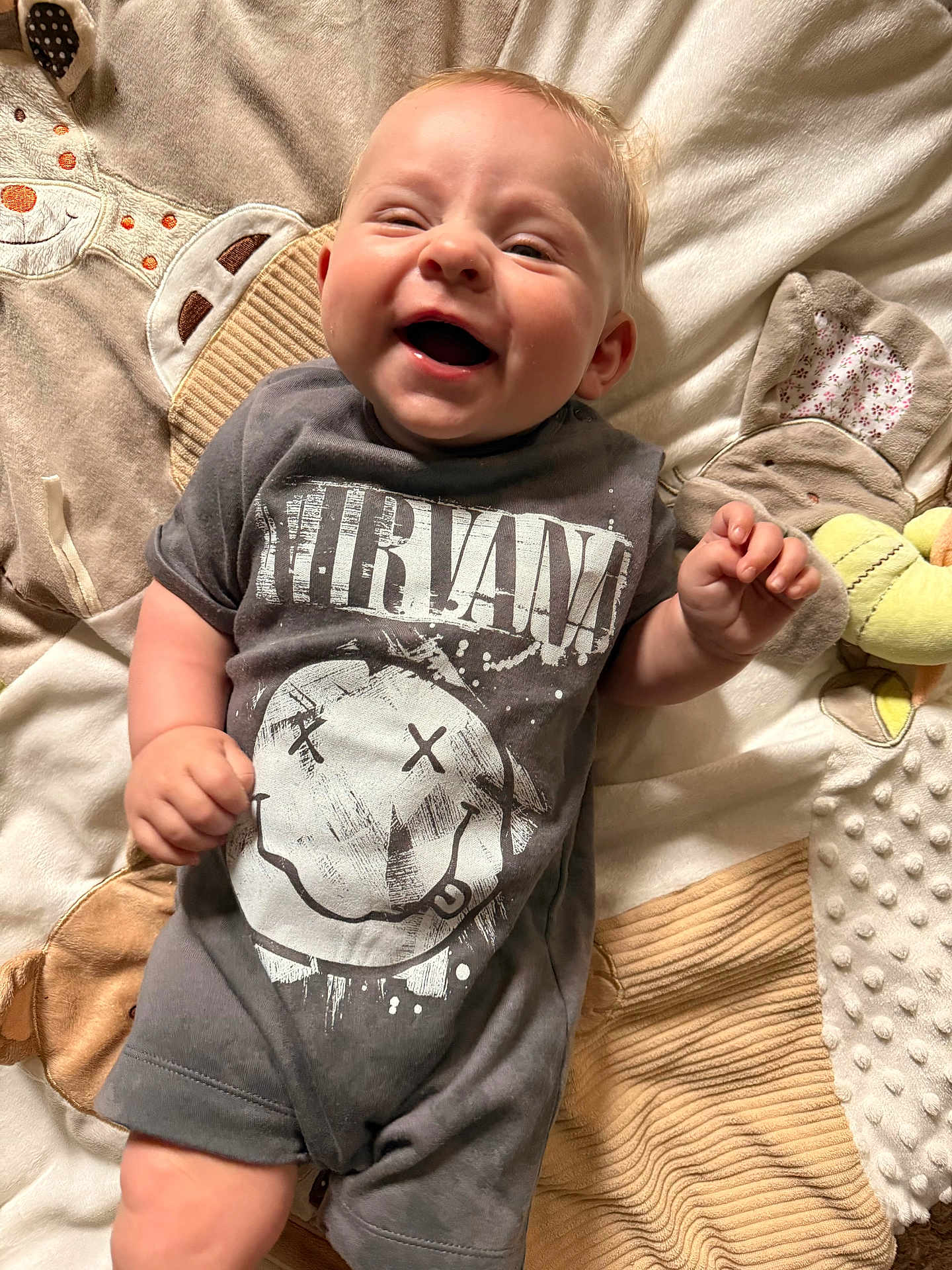 Autumn is registered to the contest to win money with this photo: baby, smiling, laughing, infant, nirvana_tshirt, clothing, blanket, soft_toy, cute, happy, lying_down, child, indoor, face, young_child, toddler, person, apparel, closeup, expression