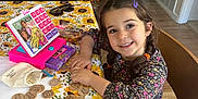 Jolie Elias is registered to the contest to win money with this photo: barbie, braids, casual_clothing, chair, child, daylight, furniture, girl, happy, home, indoor, living_room, match_game, play, portrait, smiling, sunflower_tablecloth, table, toy, wooden_tokens